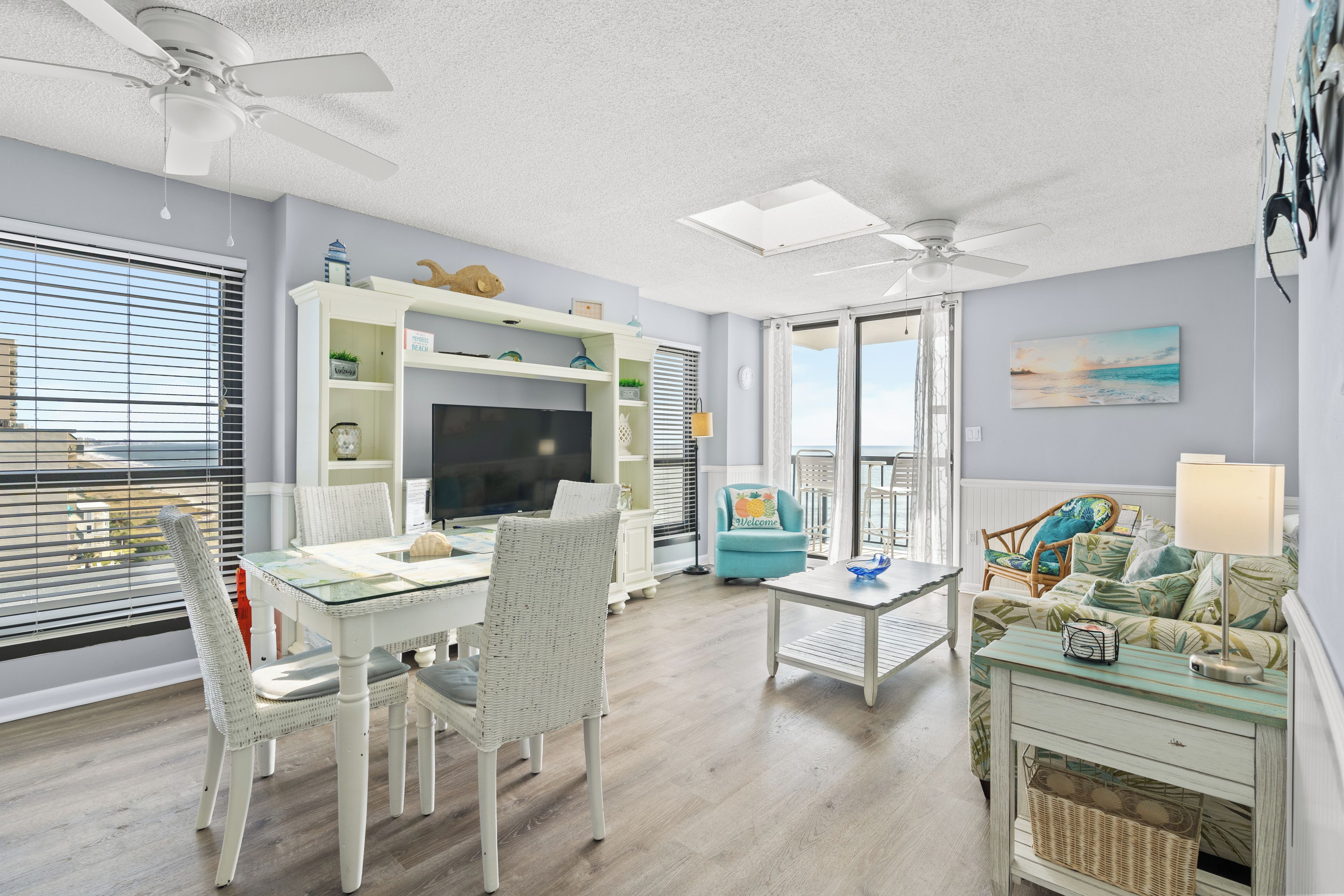 A living room with a glass-top dining table, white wicker chairs, a teal armchair, a white sofa with a patterned throw, and a ceiling fan.