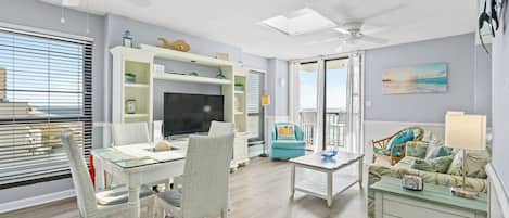 A living room with a glass-top dining table, white wicker chairs, a teal armchair, a white sofa with a patterned throw, and a ceiling fan.