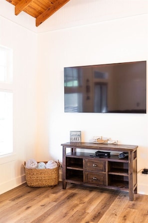 A room with a wooden floor, a flat-screen TV mounted on the wall, a dark wooden console table with drawers, a wicker basket with towels, and a sign that reads 'LOVE LAKE'. - Chalet Premiere at Signal Point (Guntersville)