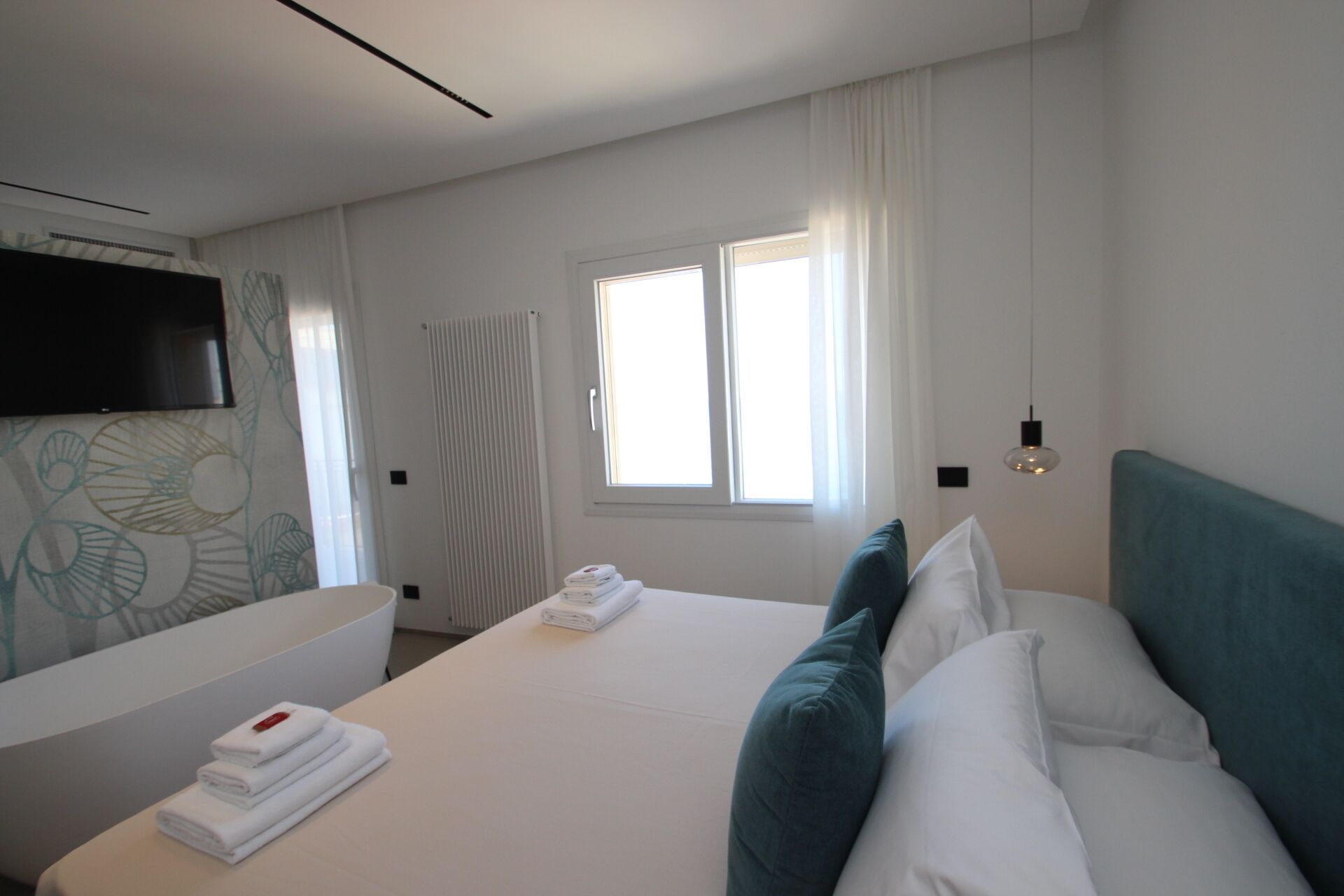 Una habitación de hotel con cama, televisor montado en la pared, ventana con cortinas y un perchero para toallas.