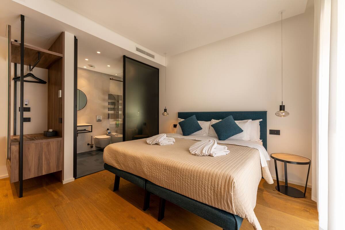 A modern bedroom with a large bed, a bedside table, a hanging light, and a view into the bathroom.