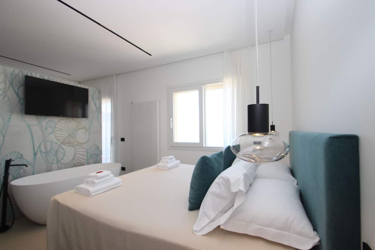 A modern hotel room with a bed, a large window, a television, and a bathroom with a bathtub.