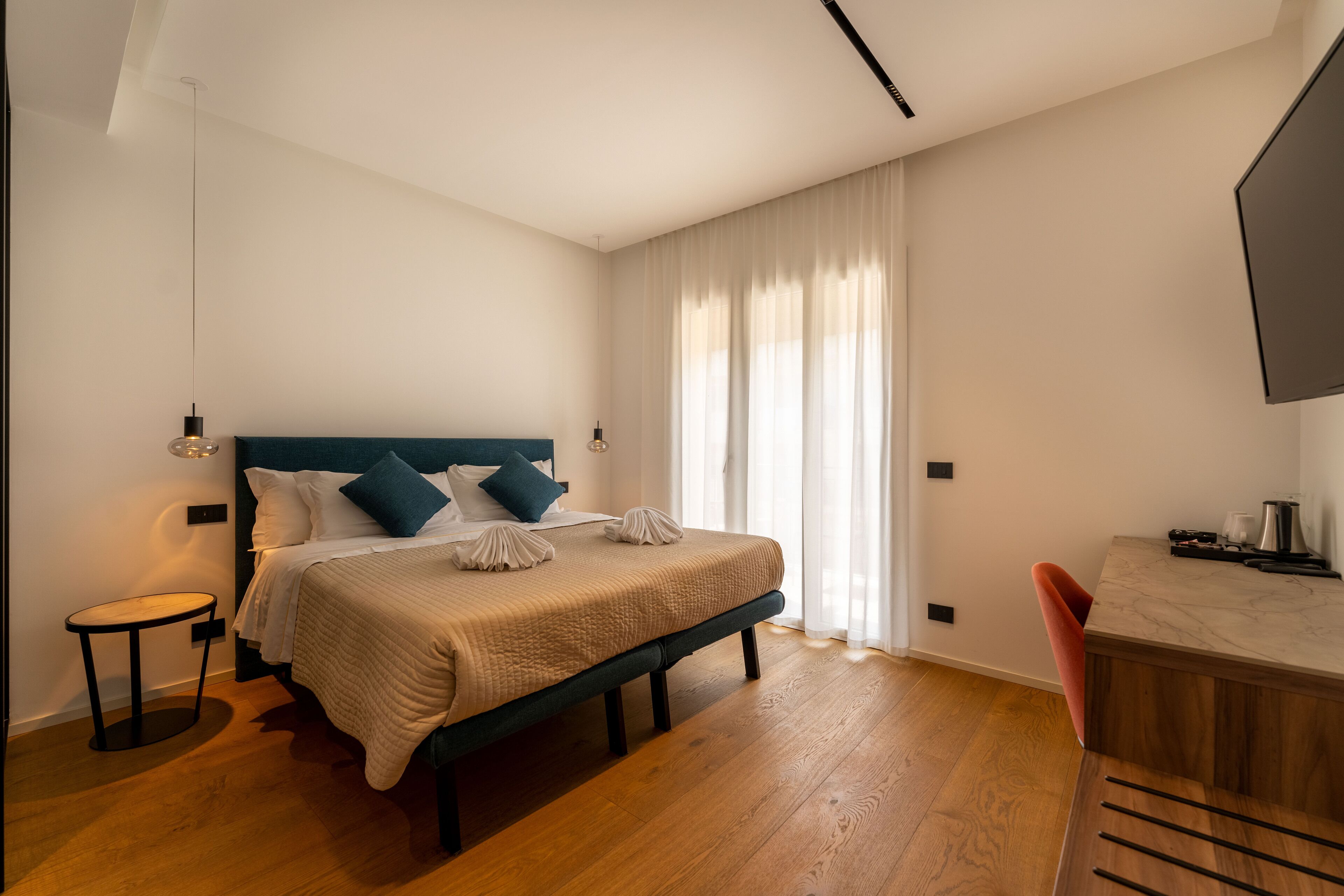 A modern hotel room with a large bed, a small side table, a desk with a chair, and a large window with curtains.