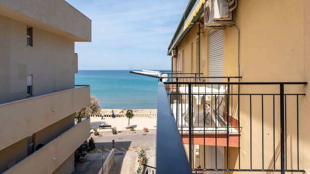 Un balcón con vistas al mar, un edificio de fachada color beige y una pequeña zona de estar al aire libre con macetas.
