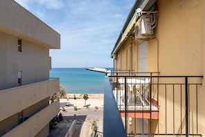 Un balcon avec vue sur la mer, un immeuble à la façade beige et un petit espace extérieur aménagé avec des plantes en pot.