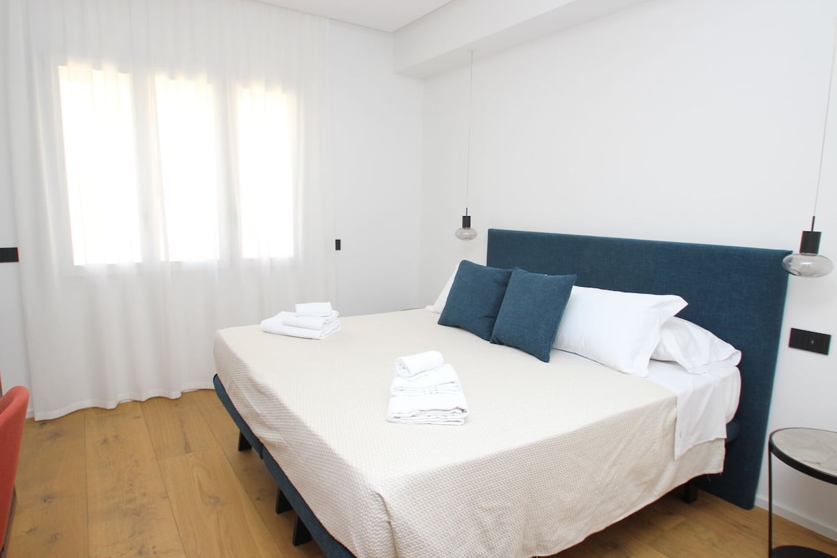 A neatly made bed with white linens and blue pillows, a wooden floor, and a small table with a chair.