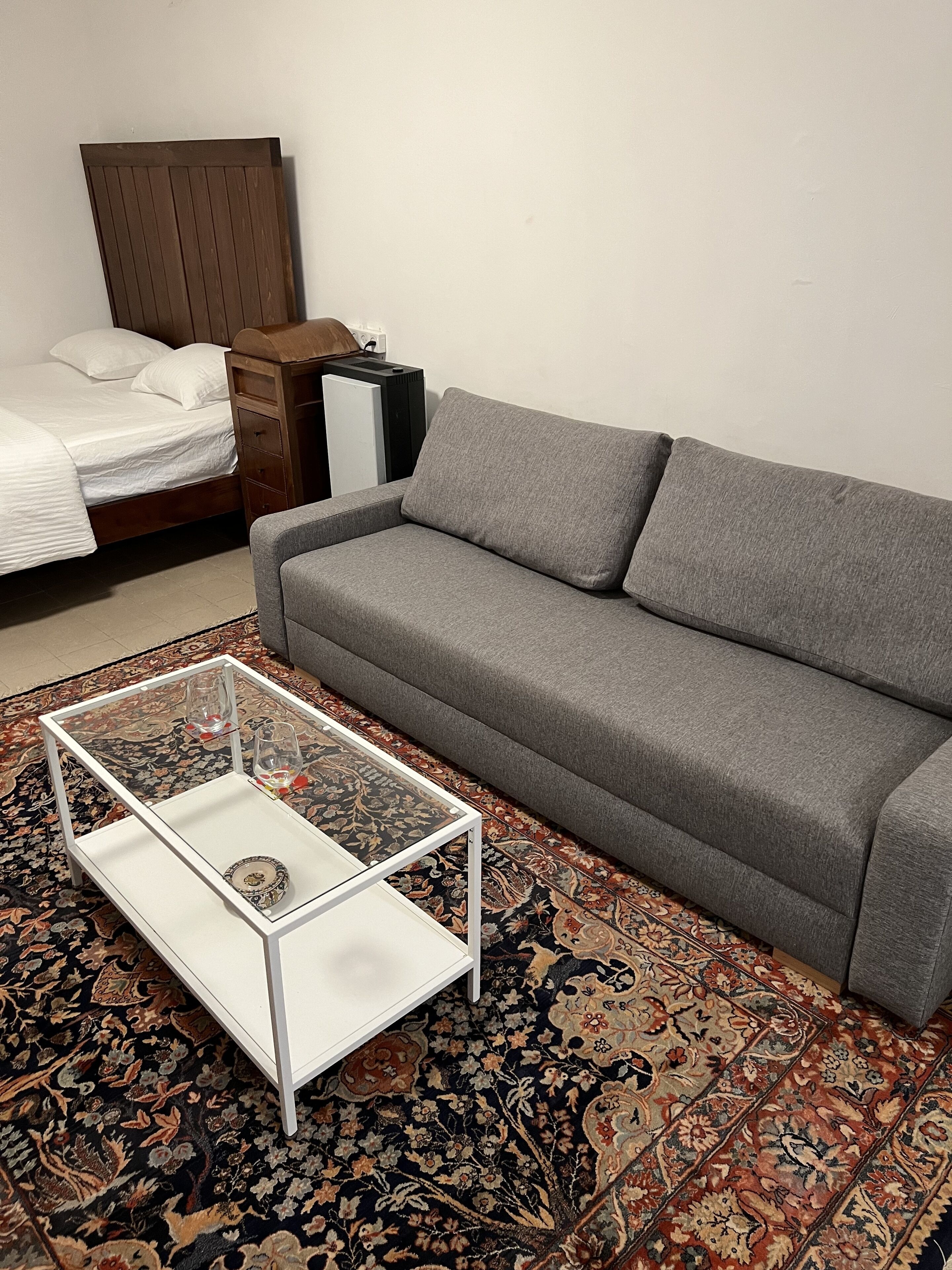A room with a grey sofa, a glass coffee table, a patterned rug, a bed with white bedding, and a wooden headboard.