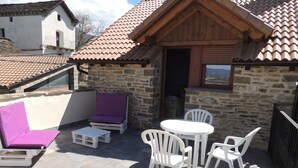 Eine Terrasse mit Steinmauer, Holzdach, einem violett gepolsterten Stuhl, einem kleinen Tisch und weißen Plastikstühlen.