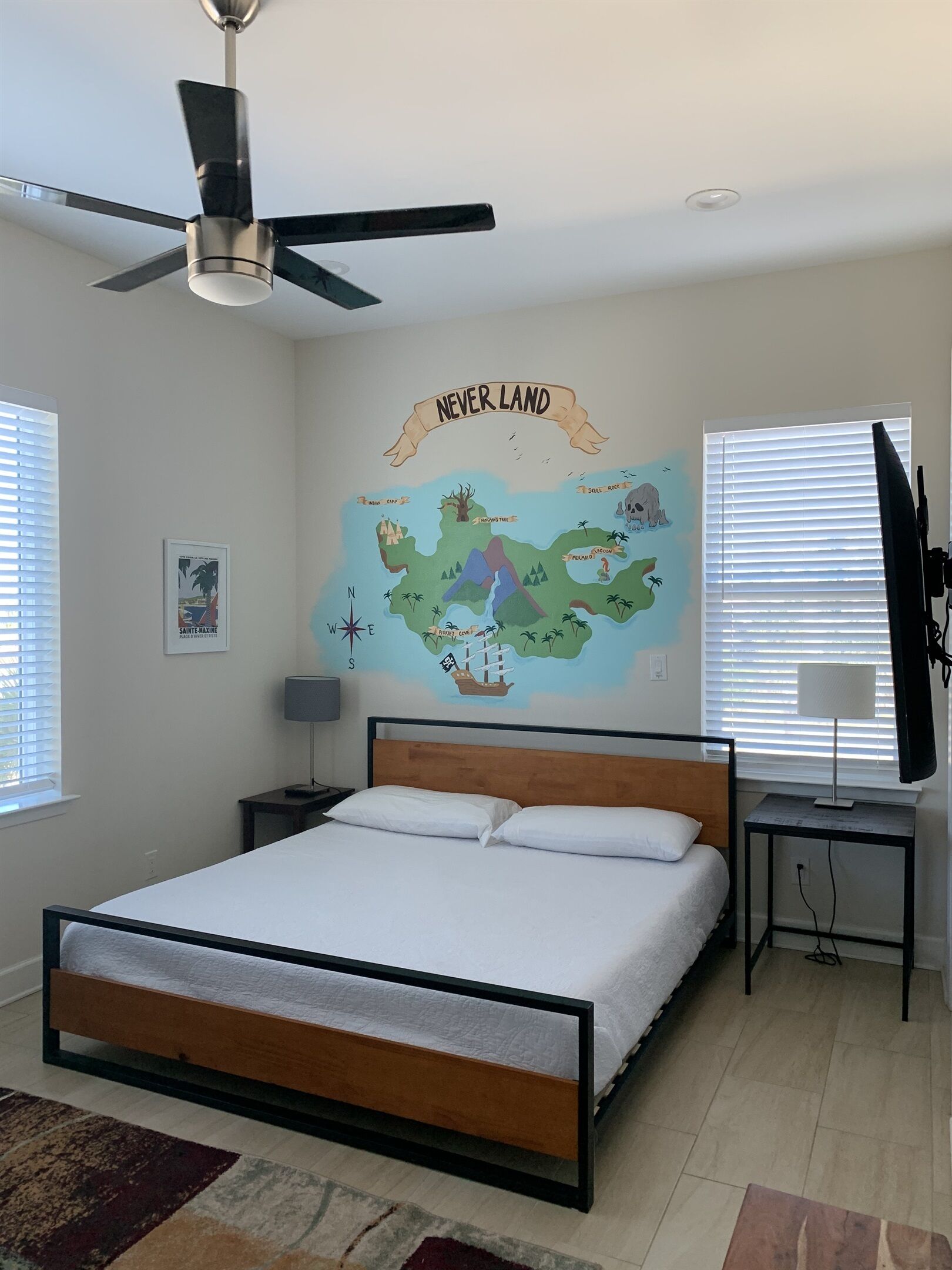 A bedroom with a bed, a ceiling fan, a map mural, a desk, and a TV.