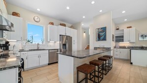 A modern kitchen with white cabinetry, a stainless steel refrigerator, a large island with bar stools, and a view of the outdoors through a window.