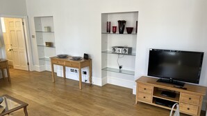 A modern living room with a wooden floor, a glass-top coffee table, a TV stand with a flat-screen TV, a bookshelf with decorative items, and a door leading to another room. - Central 1-bed Apartment in Inverness, Sleeps 4 (Inverness)