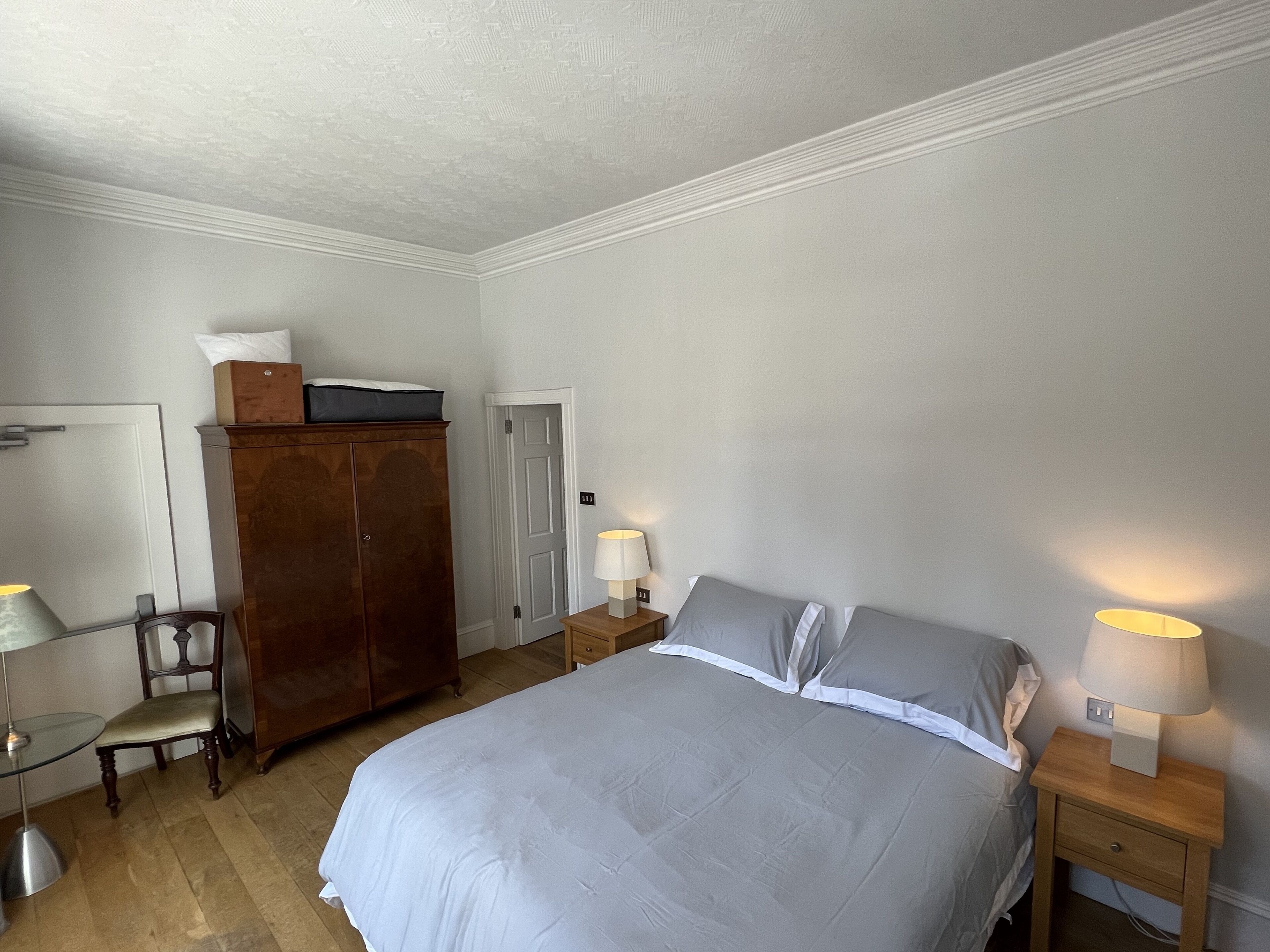 A bedroom with a bed, two bedside tables with lamps, a wooden wardrobe, a chair, and a door.