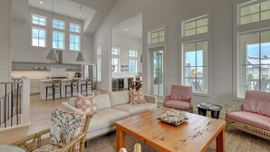 A spacious living area with a large sofa, a wooden coffee table, and wicker chairs. There is a kitchen in the background with a bar area.