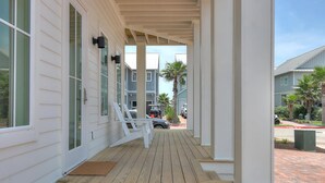 A covered porch with white railings and a wooden deck, leading to a street with parked cars and palm trees.