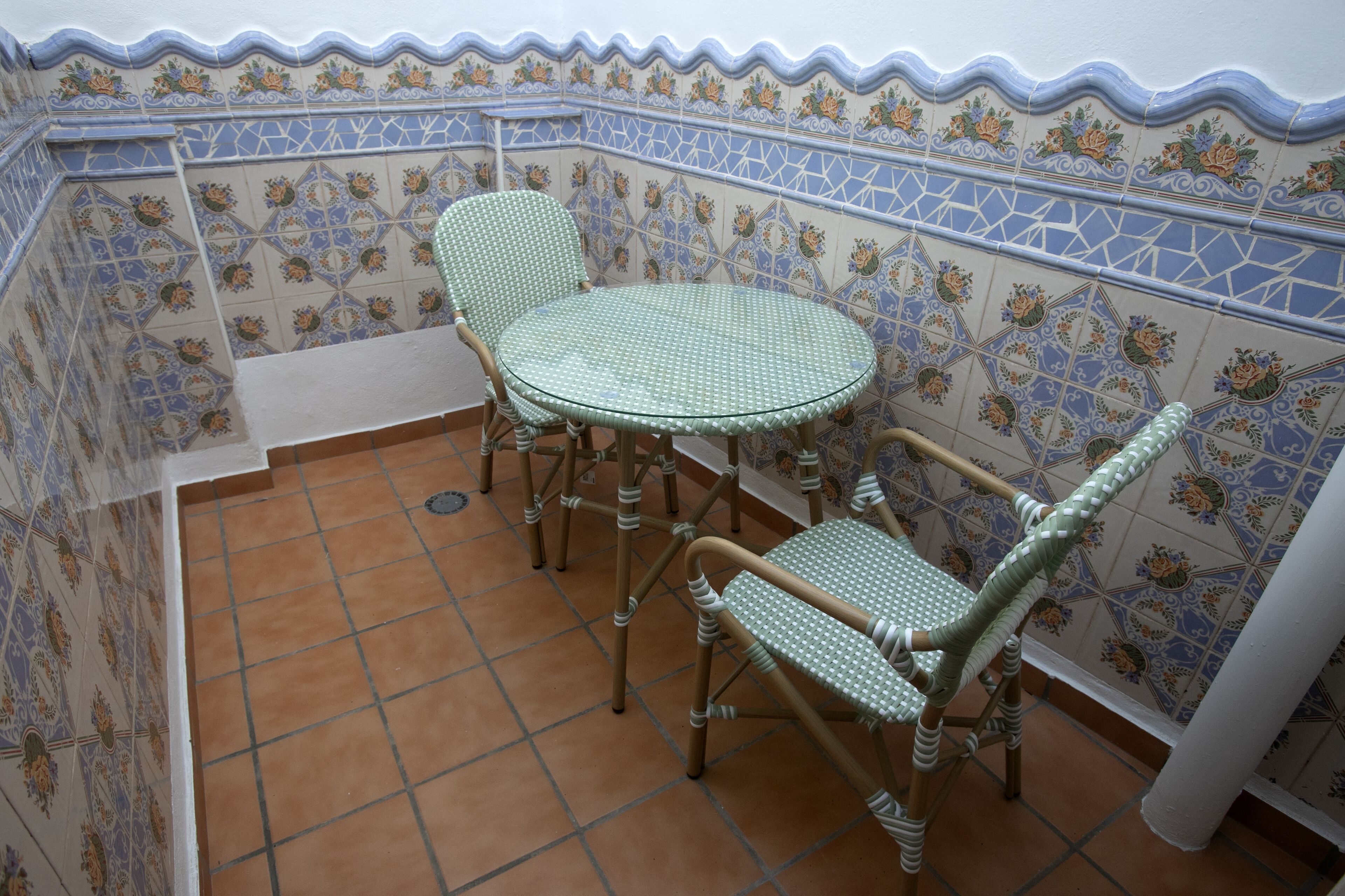 Un pequeño patio con una mesa redonda y dos sillas de mimbre, rodeado de paredes decorativas con azulejos.