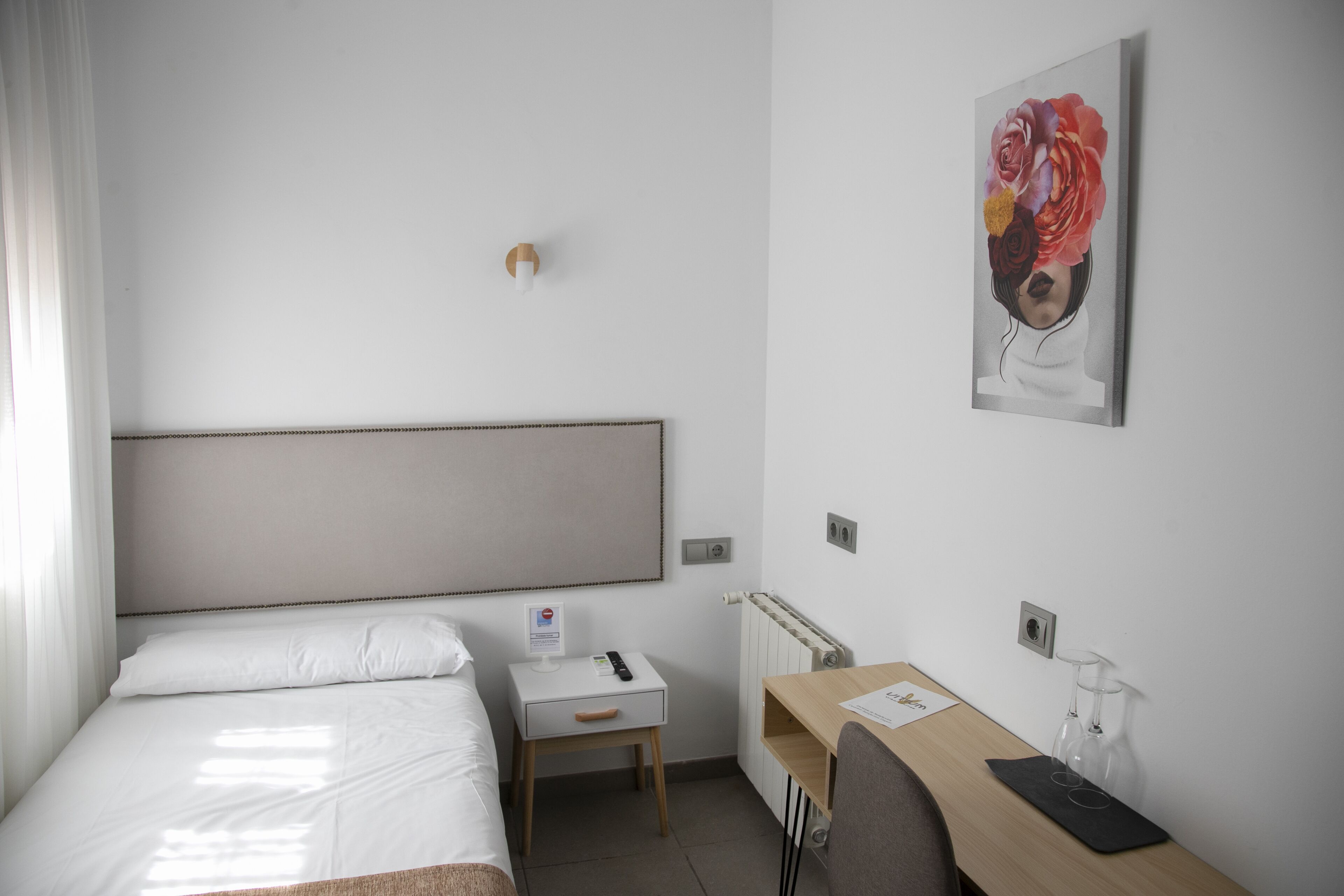 Habitación de hotel con cama, escritorio, silla, mesita pequeña, radiador y una lámpara fijada a la pared.