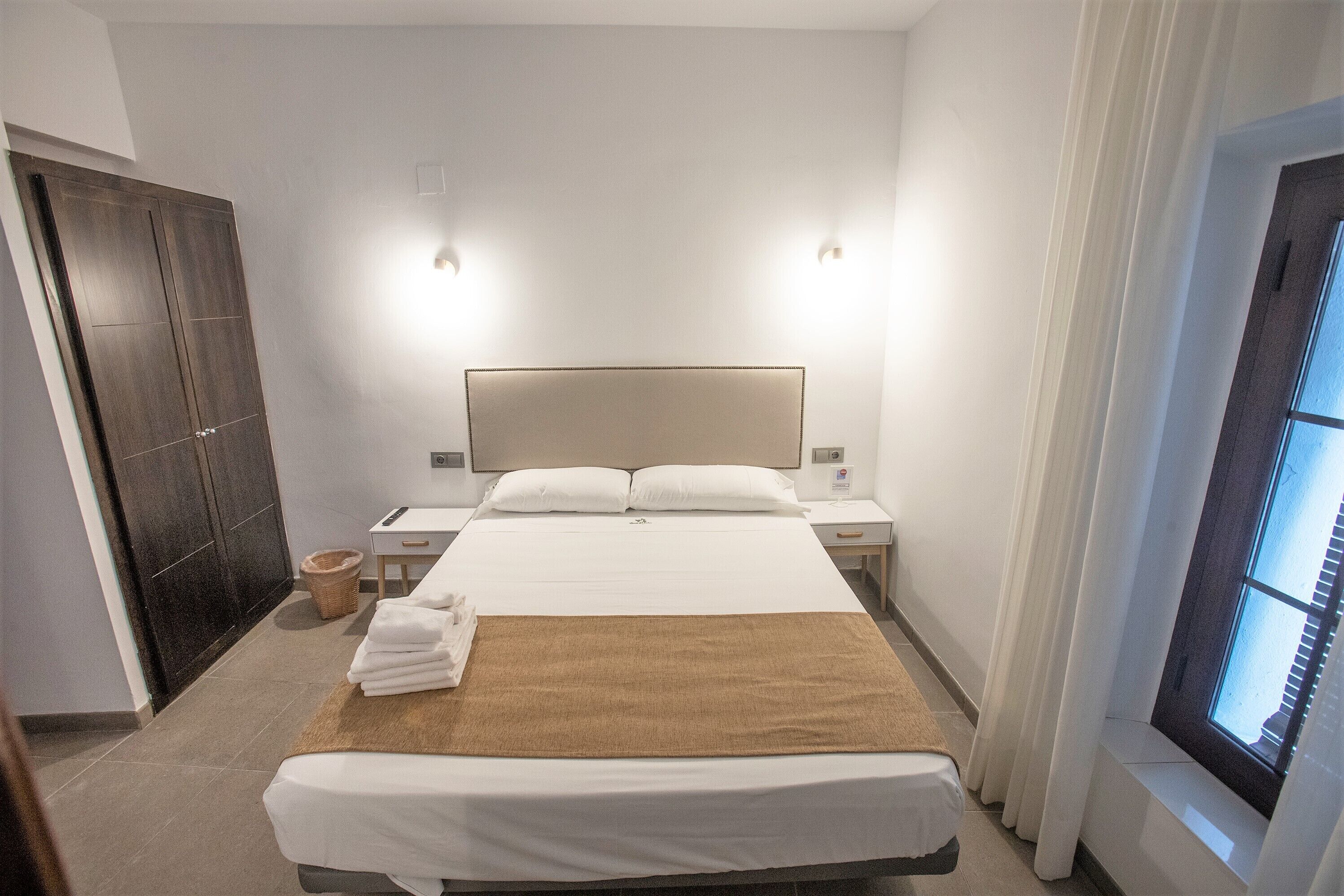 Una habitación de hotel con una cama grande, dos mesitas de noche, un armario y una ventana con cortinas.