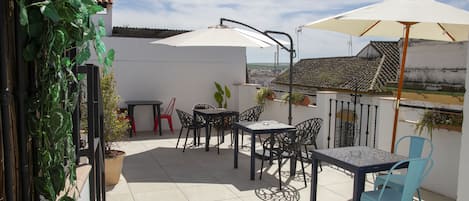 En udendørs terrasse med fliset gulv, et bord med stole og en stor parasol, der giver skygge.