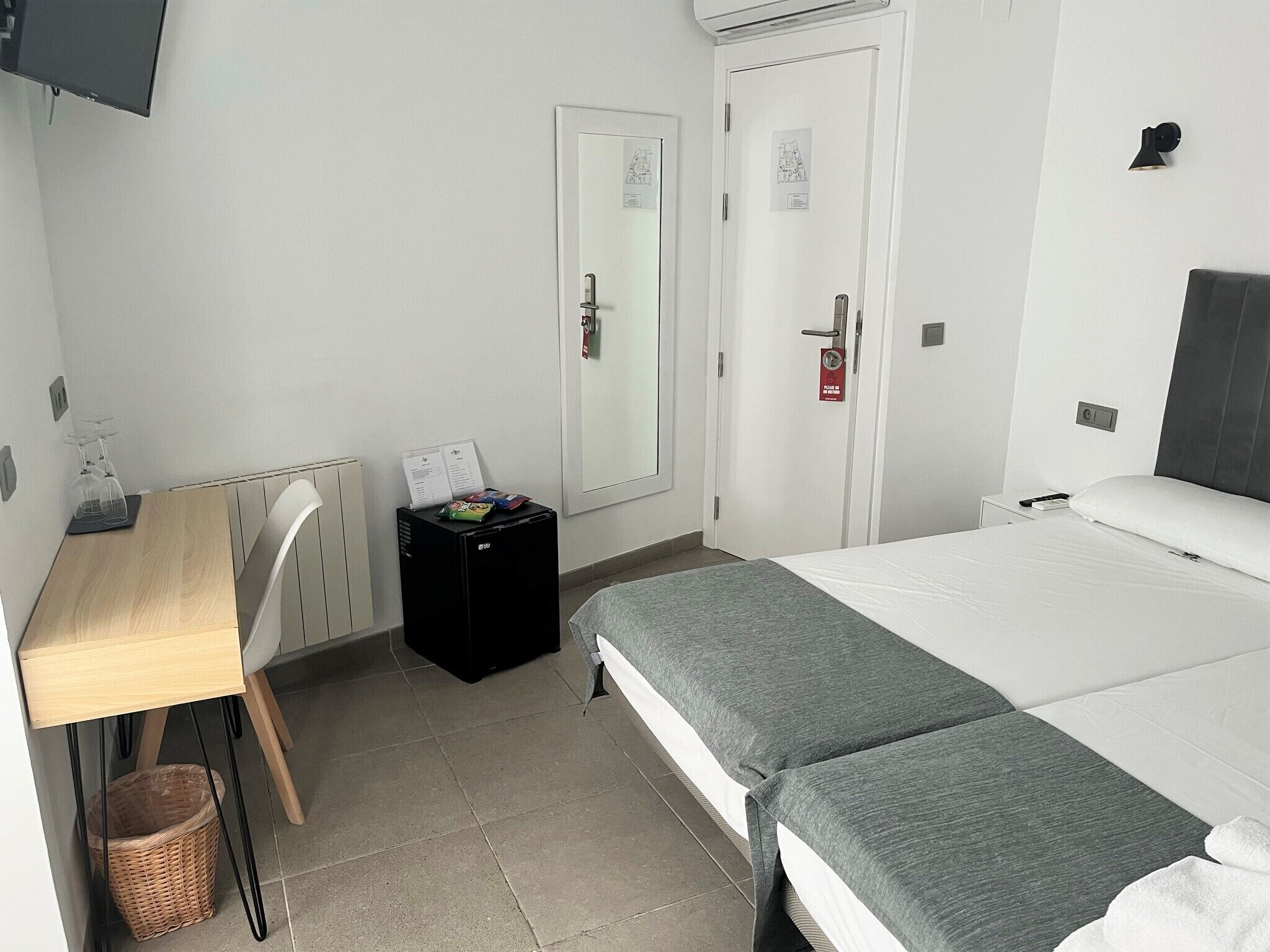Hotellihuone, jossa on sänky, työpöytä, tuoli, pieni musta kaappi, rottinkikori ja valkoinen ovi, jossa on palovaroitin.
