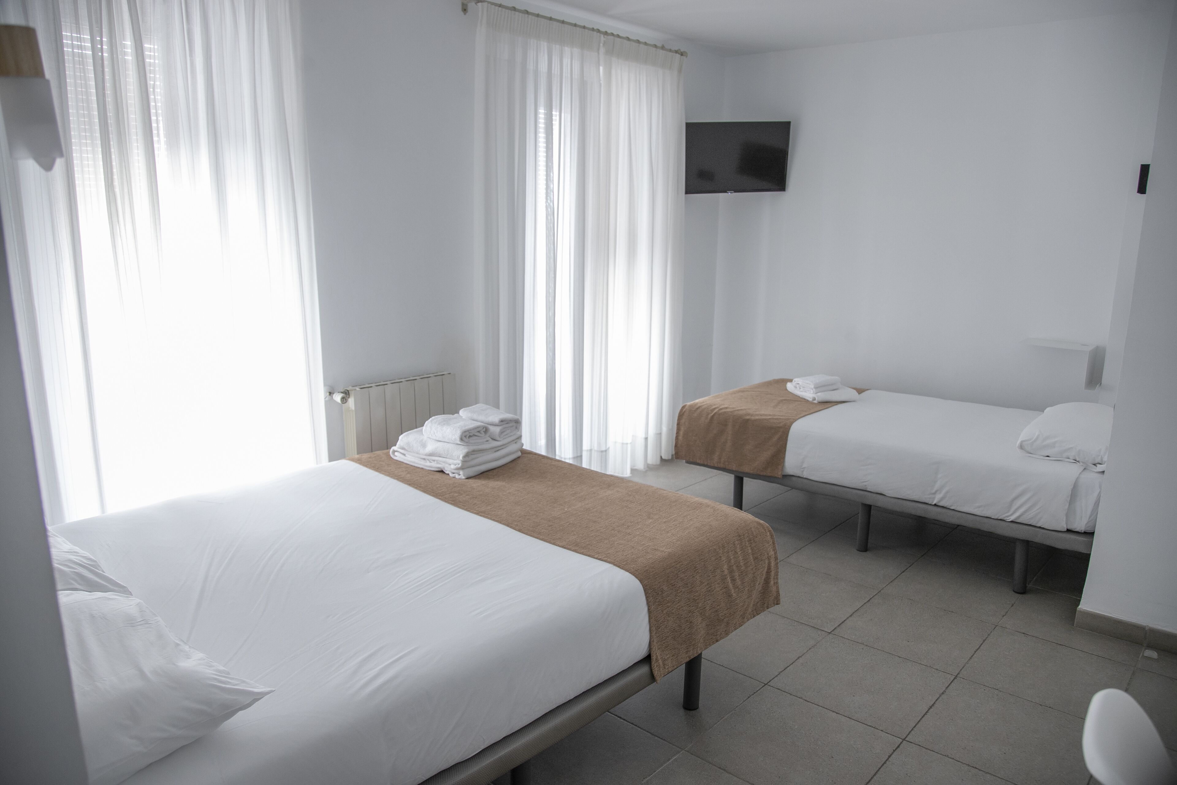 A hotel room with two single beds, a TV mounted on the wall, and a window with sheer curtains.