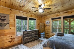 A wooden cabin bedroom with a bed, dresser, and windows offering views of the forest.