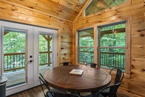 A wooden interior with a round table and chairs, large windows, and a view of greenery outside.