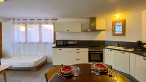 Eine kompakte Küche mit Mikrowelle, Backofen und Spüle, ein Esstisch gedeckt mit roten Tellern und Gläsern, und ein Bett im angrenzenden Raum.