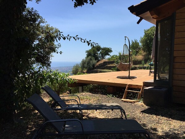 Terraza de madera con dos sillones reclinables y una silla colgante, con vistas a un paisaje pintoresco.