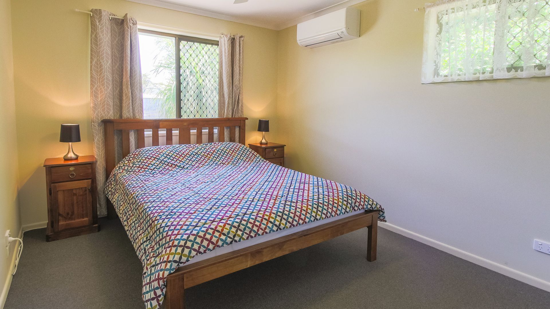 Une chambre avec un lit en bois, deux tables de chevet avec des lampes, une fenêtre avec des rideaux et un appareil de climatisation fixé au mur.