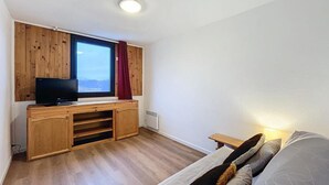 Una habitación compacta con un mueble de entretenimiento de madera, televisor, ventana con cortinas, radiador y cama con almohadas.