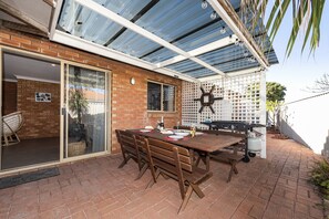 Eine Terrasse im Freien mit Ziegelwand, Glastür zu einem überdachten Bereich, Holztisch mit Stühlen und einem Grill.