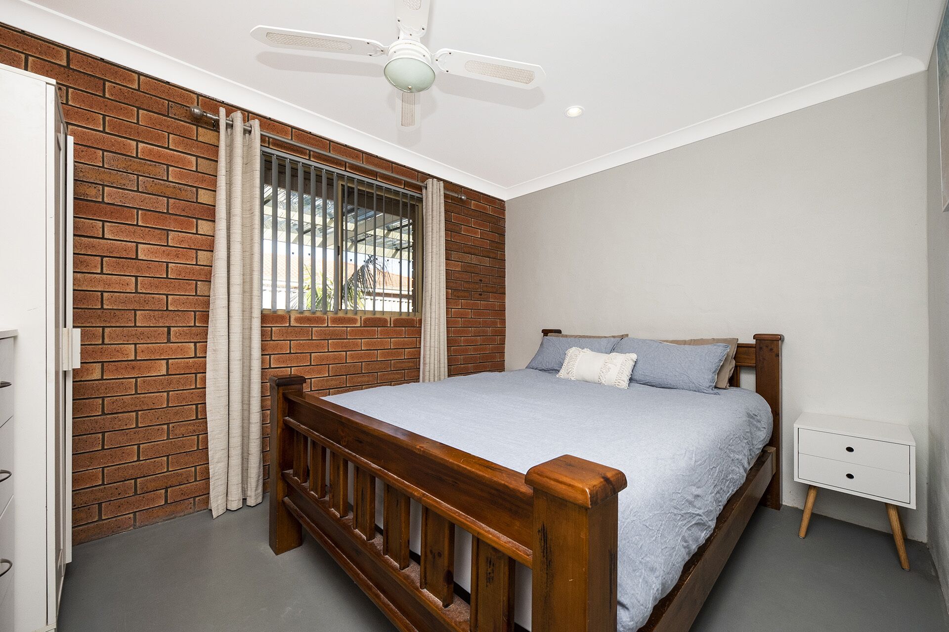 Un dormitorio con pared de ladrillo, ventilador de techo, ventana con rejas, cama con cabecero de madera y mesita de noche.