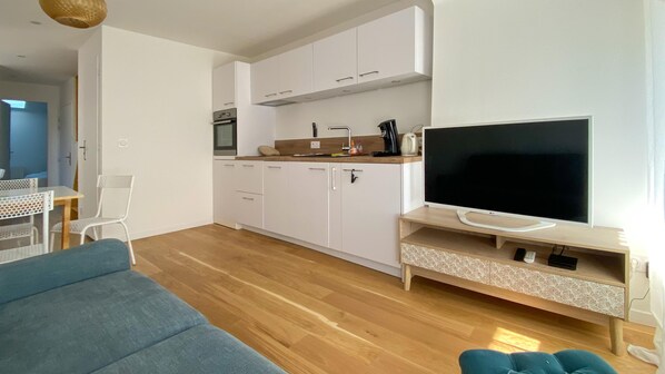 Un salon moderne avec un canapé, un téléviseur posé sur un meuble en bois et un coin cuisine doté de meubles blancs et d’une cuisinière.