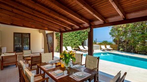 A covered outdoor dining area with a pool view, wooden furniture, and a table set for a meal. - 3 Roses Residence in Afandou (Αφάντου)