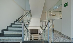 A modern staircase with glass railings and marble steps, leading to a landing with a white wall and a green sign indicating the floor level. - Itsy Hotels Vijaya Comforts, with BAR & restaurant (Somvarpet)