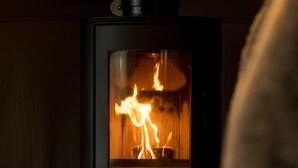 A wood-burning stove with visible flames inside, set in a room with a bed in the foreground. - Hever Luxury Dome (Hoath)