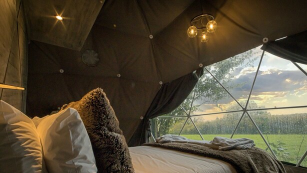 A cozy tented accommodation with a bed, pillows, and a view of a green landscape through a large window. - Hever Luxury Dome (Hoath)