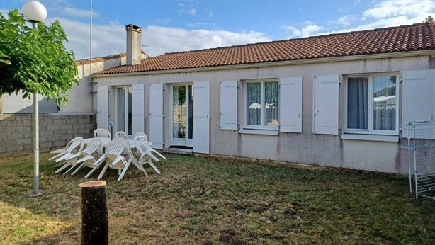 Un bâtiment doté d’un toit en tuiles, de fenêtres à cadre blanc et d’un petit espace extérieur aménagé avec des chaises en plastique blanches.