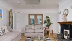 Una sala de estar con un sofá, una mesa redonda de mimbre, una chimenea y un ventanal con contraventanas de madera.