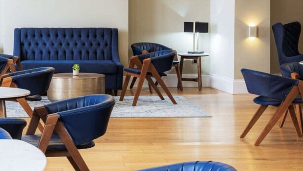 Un área de descanso moderna con un sofá azul, sillas de madera y una mesa de centro redonda.