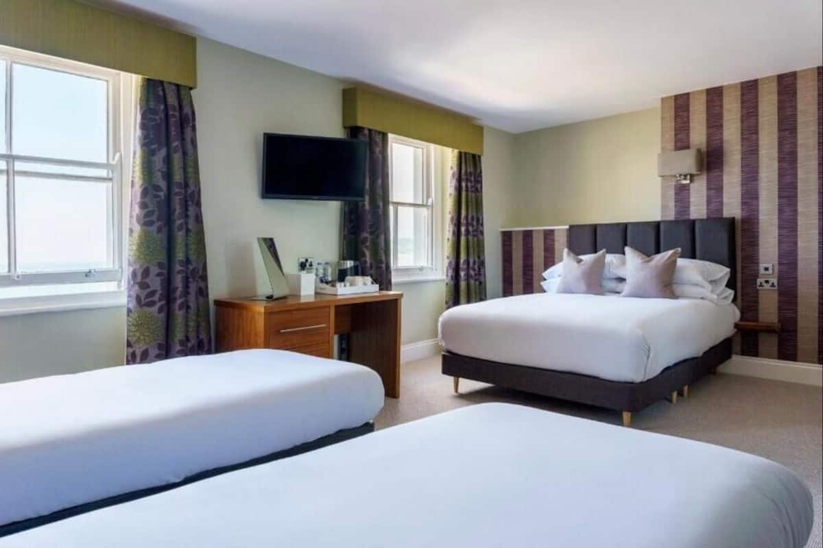 A hotel room with two single beds, a desk with a chair, a flat-screen TV mounted on the wall, and a window with curtains.