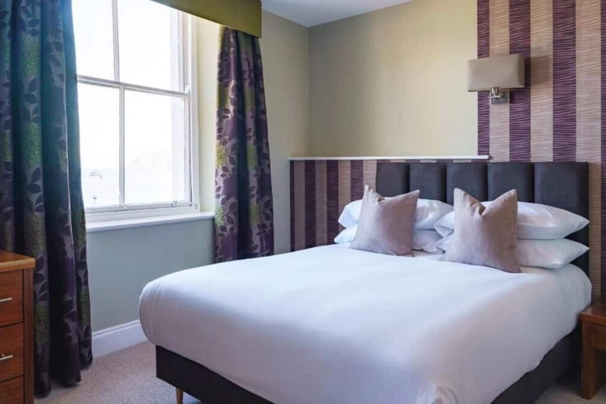 A neatly made bed with white linens and multiple pillows, a bedside lamp, and a window with patterned curtains.