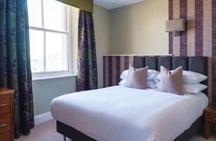 Una cama bien tendida con sábanas blancas y varias almohadas, una lámpara de noche y una ventana con cortinas estampadas.