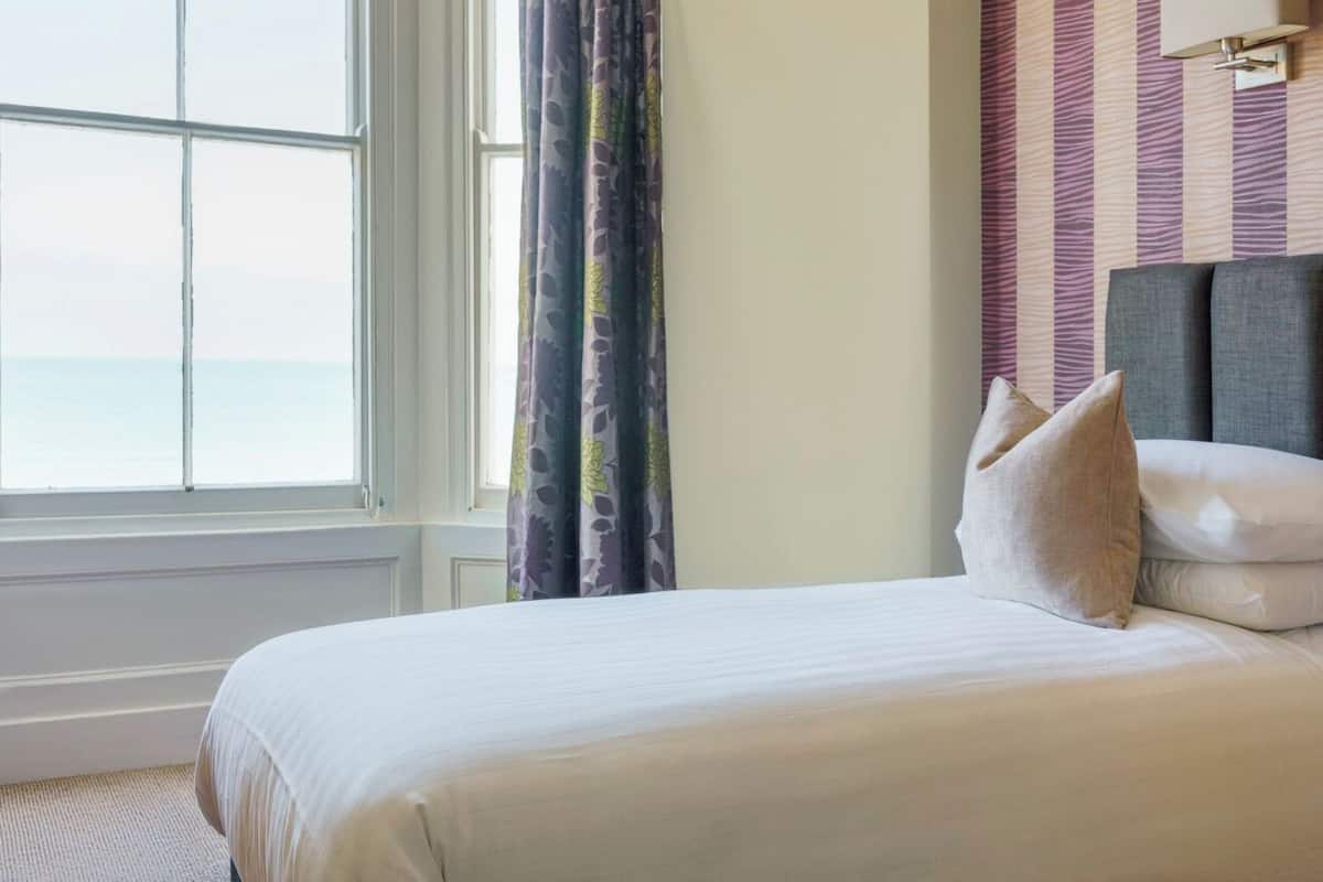 A neatly made bed with white linens, a window with a view of the sea, and a striped wall behind the bed.
