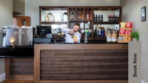 Zona de bar con un cantinero preparando una bebida, un refrigerador y estantes con botellas y aperitivos.