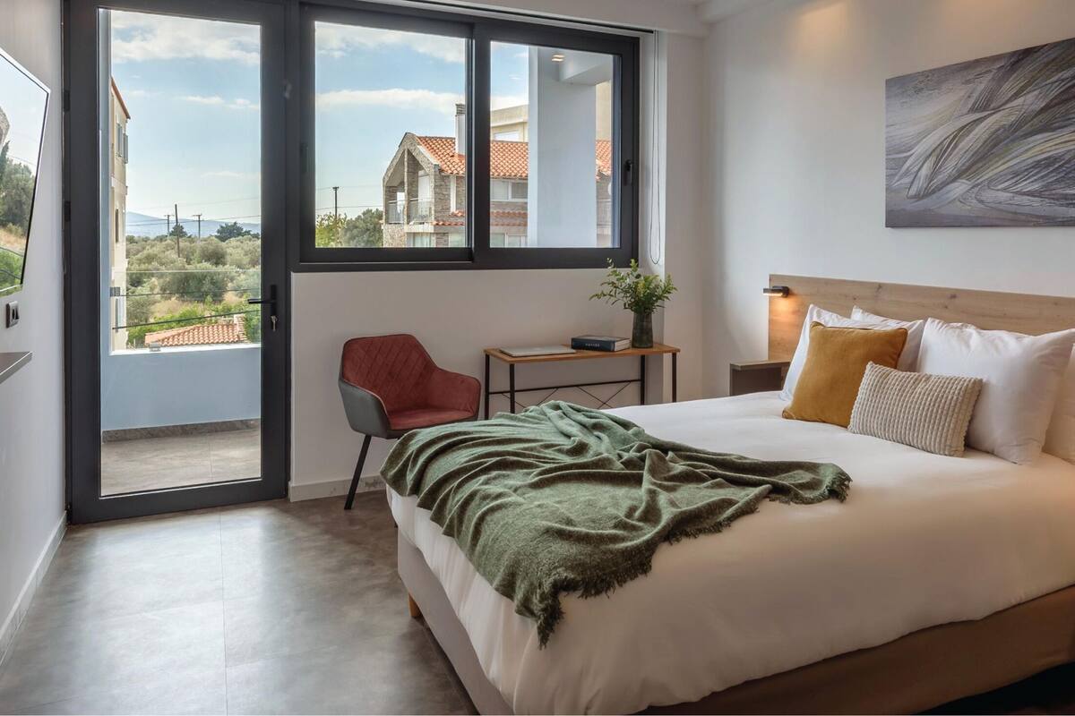 A bedroom with a large bed, a wooden headboard, a bedside table with a lamp, a chair, and a view of the outside through a sliding door.