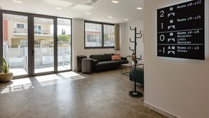 A hotel reception area with a modern design, including a reception desk, a seating area with a sofa, and a coat rack. There is a large window providing a view of the outside. A sign on the wall indicates room numbers and amenities. - Zed Smart Property by Airstay (Spata-Artemida)