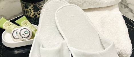 A bathroom with a marble wall, a pair of white slippers, a rolled white towel, a black and green toiletry bag, and two green toiletry tubes.