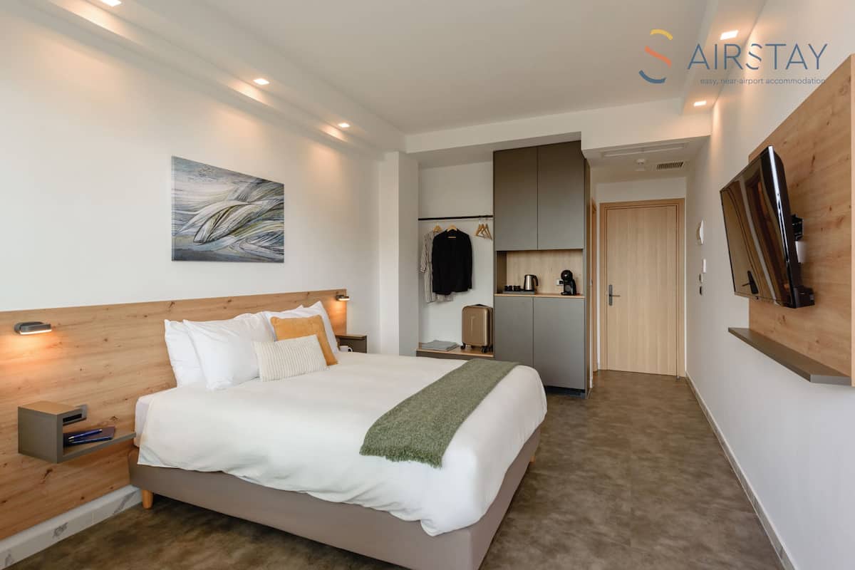 A modern hotel room with a large bed, a wooden headboard, a painting on the wall, a kitchenette, and a TV mounted on the wall.