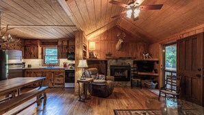 A rustic cabin interior with wooden walls, a stone fireplace, a dining area with a bench, a television, and a ceiling fan. - Twin Creeks Cabin - RIVER FRONT! *Hot tub* 4 acres! Pets OK (Deep Gap)